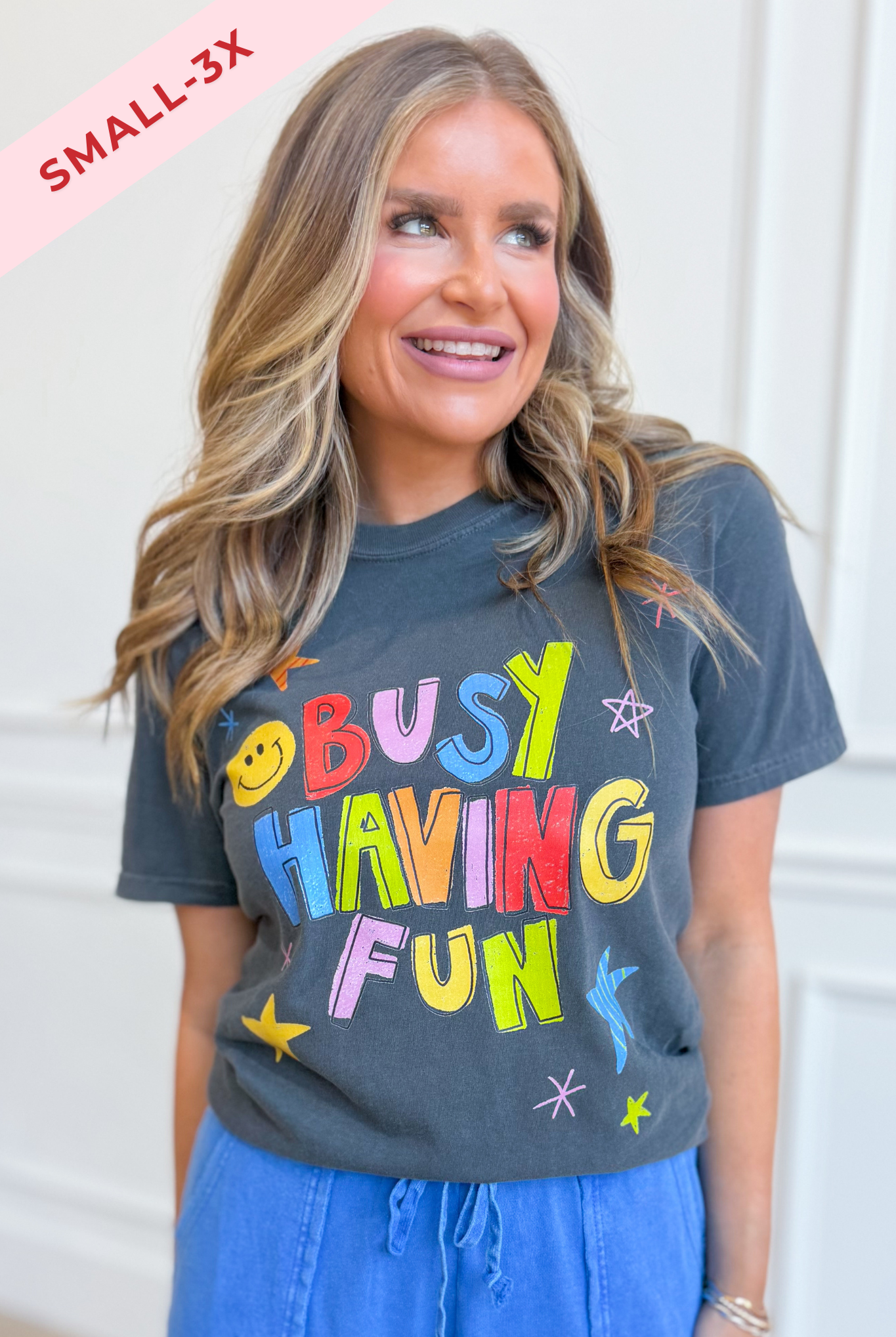 A woman wearing a dark charcoal gray graphic t-shirt with the colorful text 'BUSY HAVING FUN' and blue drawstring pants. A pink diagonal banner in the top left corner reads 'SMALL-3X'.