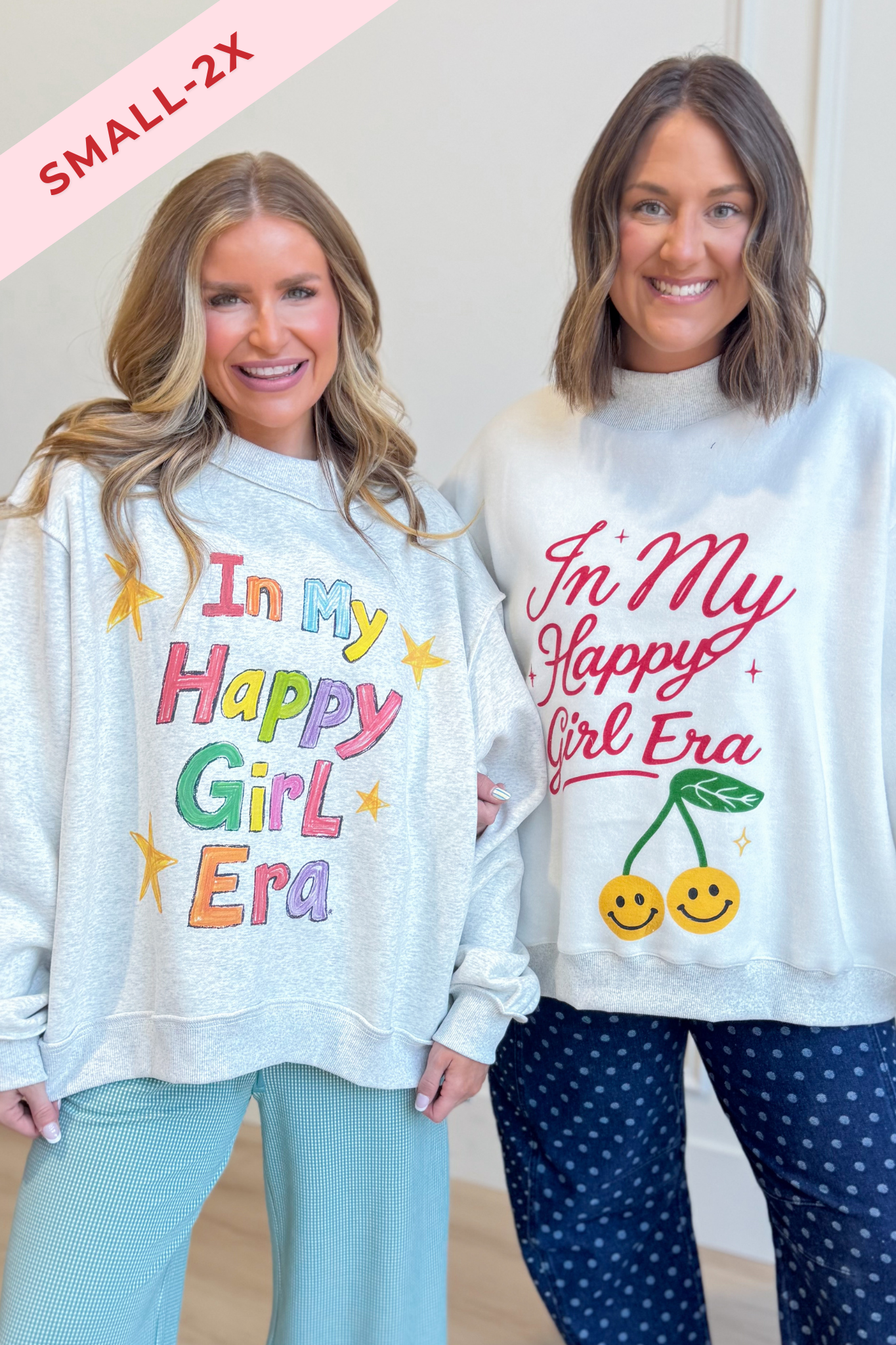 Two women modeling matching oversized sweatshirts with the phrase 'In My Happy Girl Era' in colorful lettering. The sweatshirt on the left is light gray with multicolor text and star graphics, paired with light blue pants.