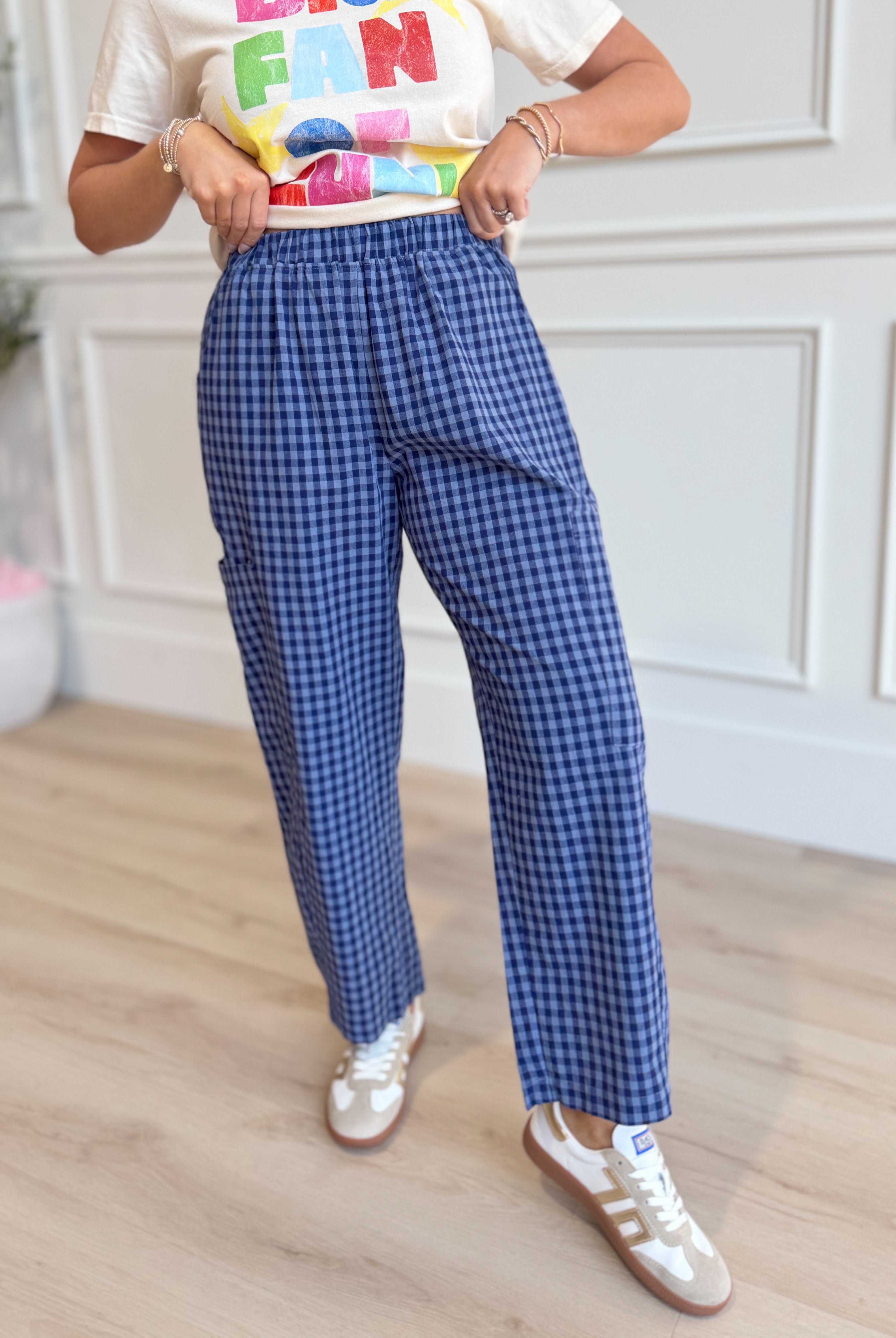 A person wearing blue and white gingham check wide-leg pants, a white graphic t-shirt, and white sneakers, standing in a room with light wood floors and white paneled walls.