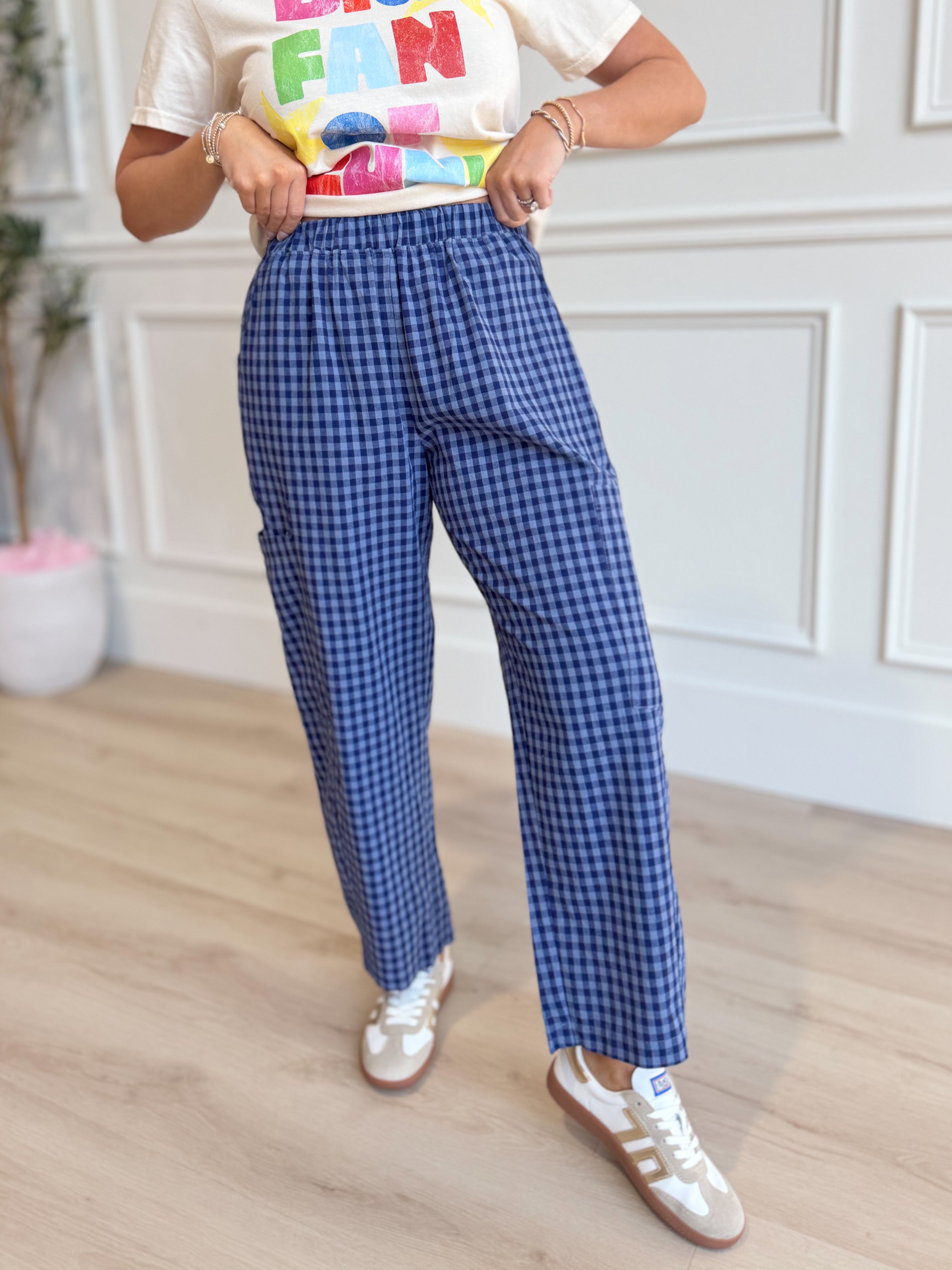 A person wearing blue and white gingham check wide-leg pants, a white graphic t-shirt, and white sneakers, standing in a room with light wood floors and white paneled walls.