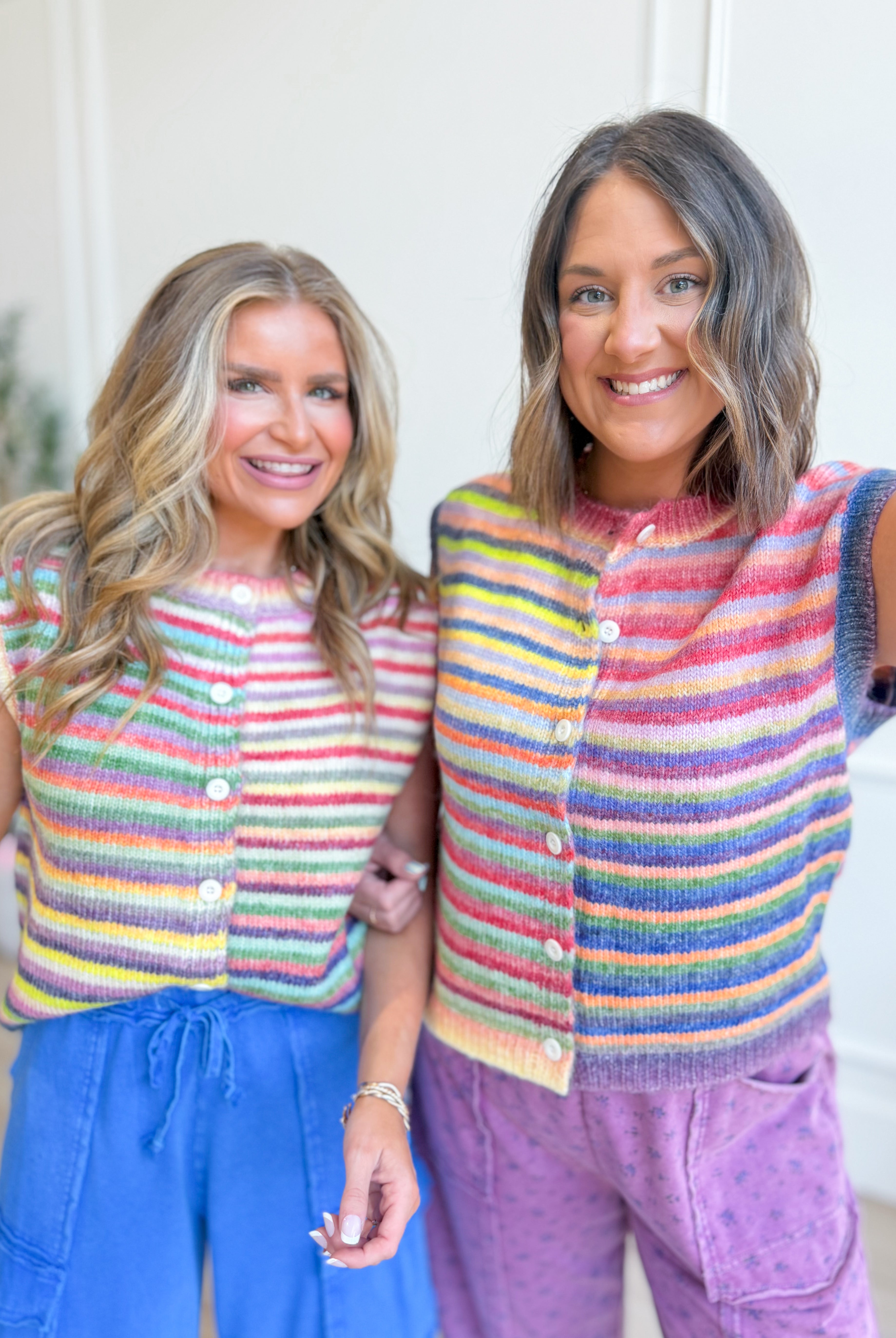 Two women smiling and wearing matching knitted button-front cardigans with horizontal rainbow stripes in pink, blue, yellow, green, and orange, paired with blue and purple lounge bottoms, standing indoors against a light neutral wall.