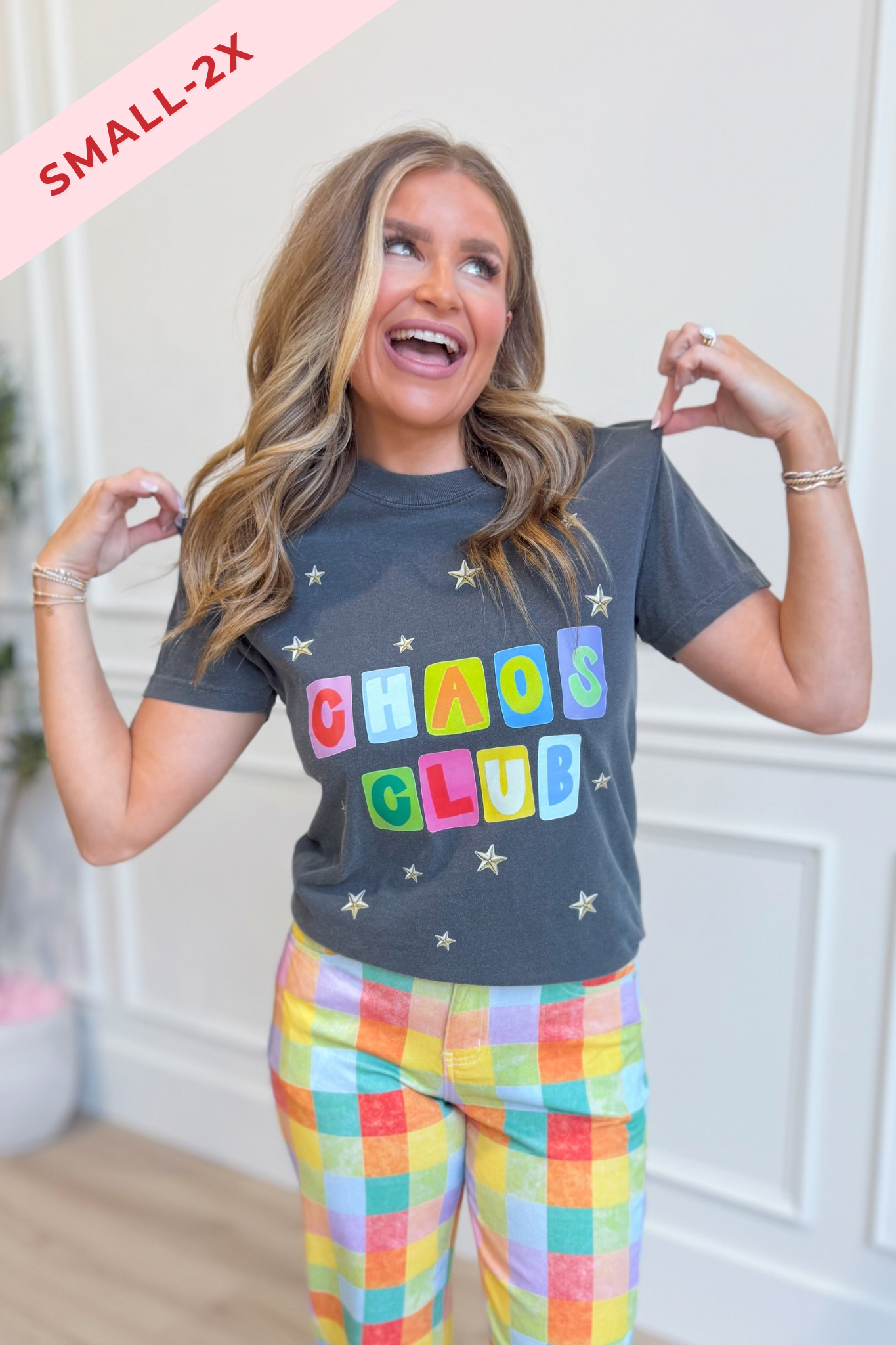A woman modeling a dark gray graphic t-shirt with 'CHAOS CLUB' printed in colorful letters, paired with pastel plaid pants. A pink diagonal banner in the top left corner reads 'SMALL-2X'.