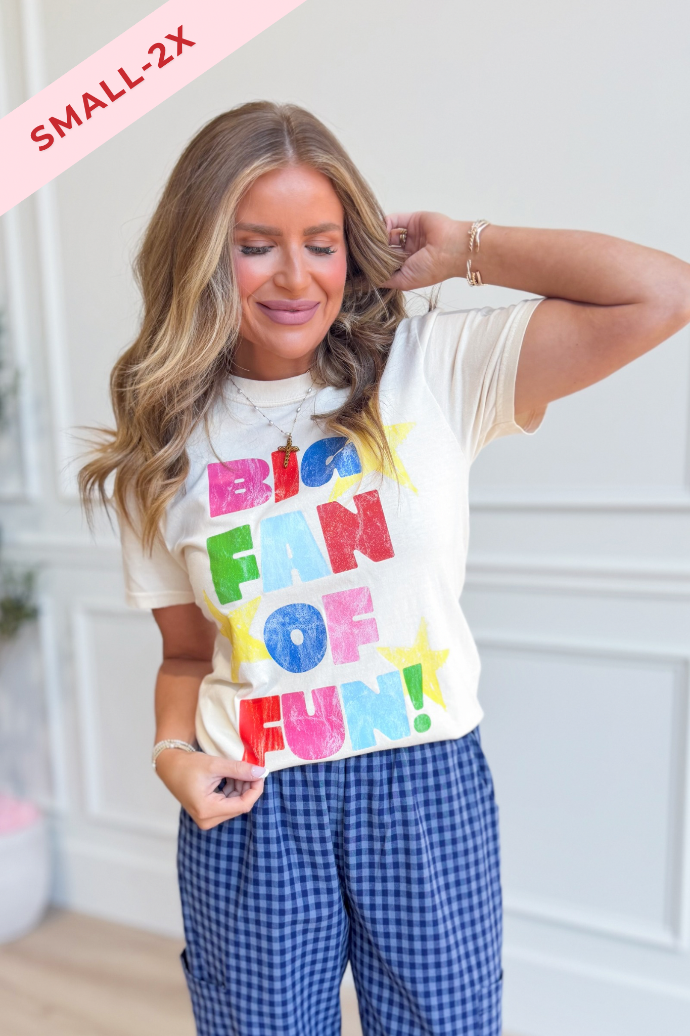 A woman wearing a cream-colored graphic t-shirt with 'BIG FAN OF FUN!' printed in colorful letters, paired with blue and white gingham pants. A pink diagonal banner in the top left corner reads 'SMALL-2X'.