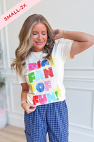 A woman wearing a cream-colored graphic t-shirt with 'BIG FAN OF FUN!' printed in colorful letters, paired with blue and white gingham pants. A pink diagonal banner in the top left corner reads 'SMALL-2X'.