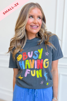 A woman wearing a dark charcoal gray graphic t-shirt with the colorful text 'BUSY HAVING FUN' and blue drawstring pants. A pink diagonal banner in the top left corner reads 'SMALL-3X'.