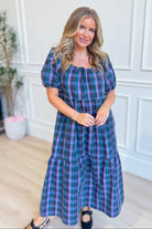 A woman modeling a short puff-sleeve midi dress with a blue, purple, and green plaid pattern, standing in a bright room with white paneled walls and light wood floors.