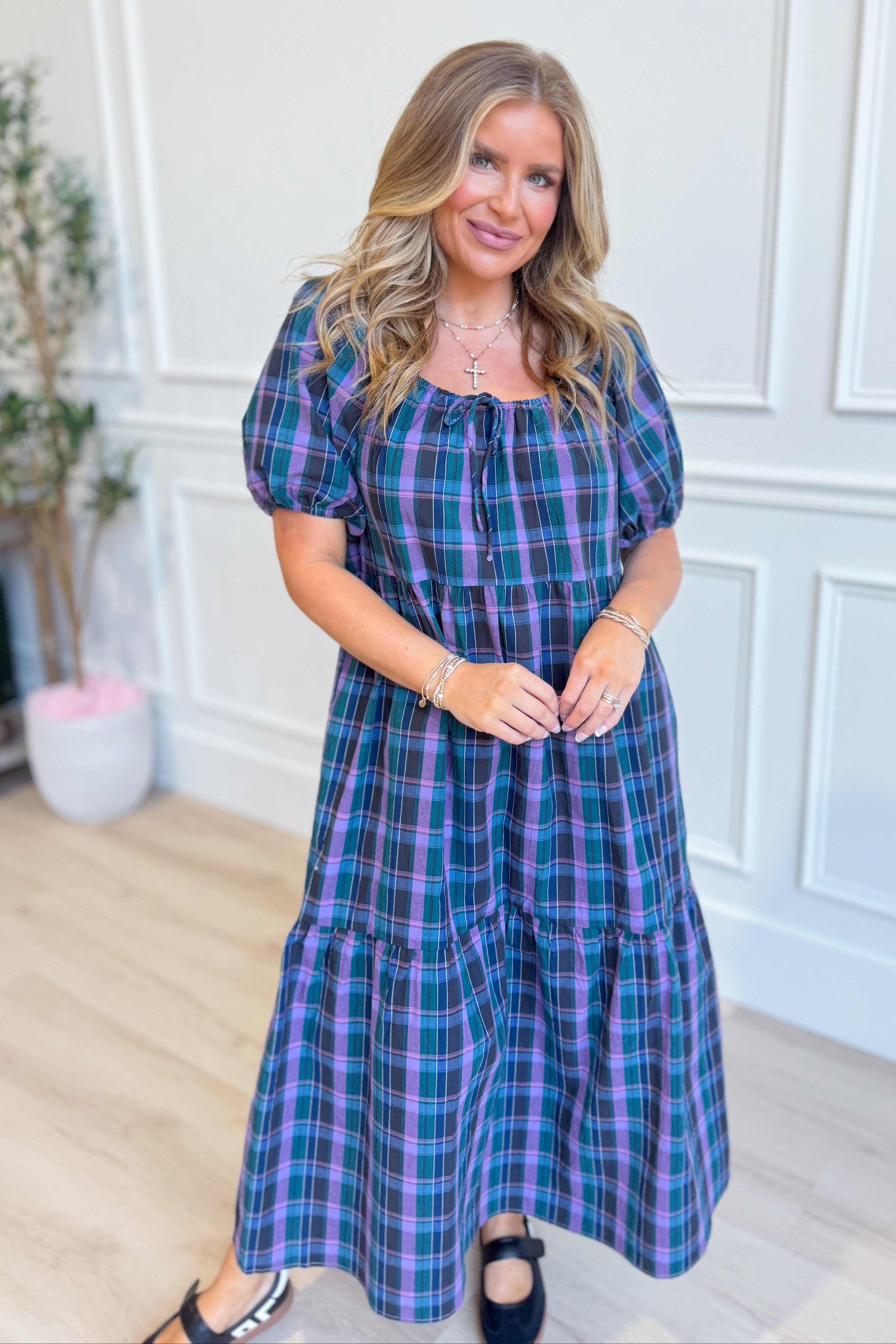 A woman modeling a short puff-sleeve midi dress with a blue, purple, and green plaid pattern, standing in a bright room with white paneled walls and light wood floors.