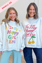 Two women modeling matching oversized sweatshirts with the phrase 'In My Happy Girl Era' in colorful lettering. The sweatshirt on the left is light gray with multicolor text and star graphics, paired with light blue pants.