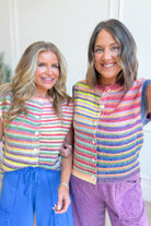 Two women smiling and wearing matching knitted button-front cardigans with horizontal rainbow stripes in pink, blue, yellow, green, and orange, paired with blue and purple lounge bottoms, standing indoors against a light neutral wall.