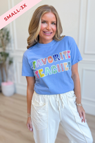 A woman wearing a light blue graphic t-shirt that reads 'FAVORITE TEACHER' in multicolored bubble letters, paired with cream-colored drawstring pants. A pink banner in the top-left corner reads 'SMALL-3X'.